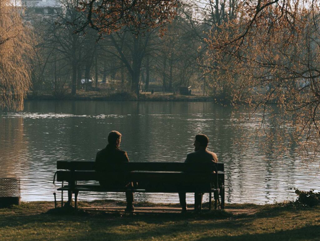 Warum uns manche Worte so schwer über die Lippen kommen: Die Kunst, Konflikte friedlich und mutig anzusprechen 2 Zwei Menschen sitzen in ein Gespräch vertieft auf einer Parkbank vor einem Teich.