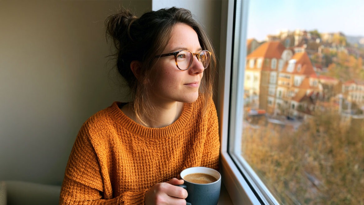 Eine nachdenkliche Frau sitzt mit einer Kaffeetasse am Fenster und blickt an einem sonnigen Morgen ins Freie.