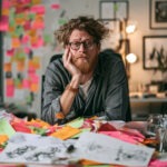Nachdenklicher Mann an einem Schreibtisch voller bunten Post-its und Skizzen als Symbol für Überforderung