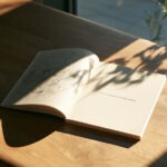 Aufgeschlagenes Notizbuch auf Holzschreibtisch mit skizzierten Zeilen links und einem klaren Satz rechts im Sonnenlicht.